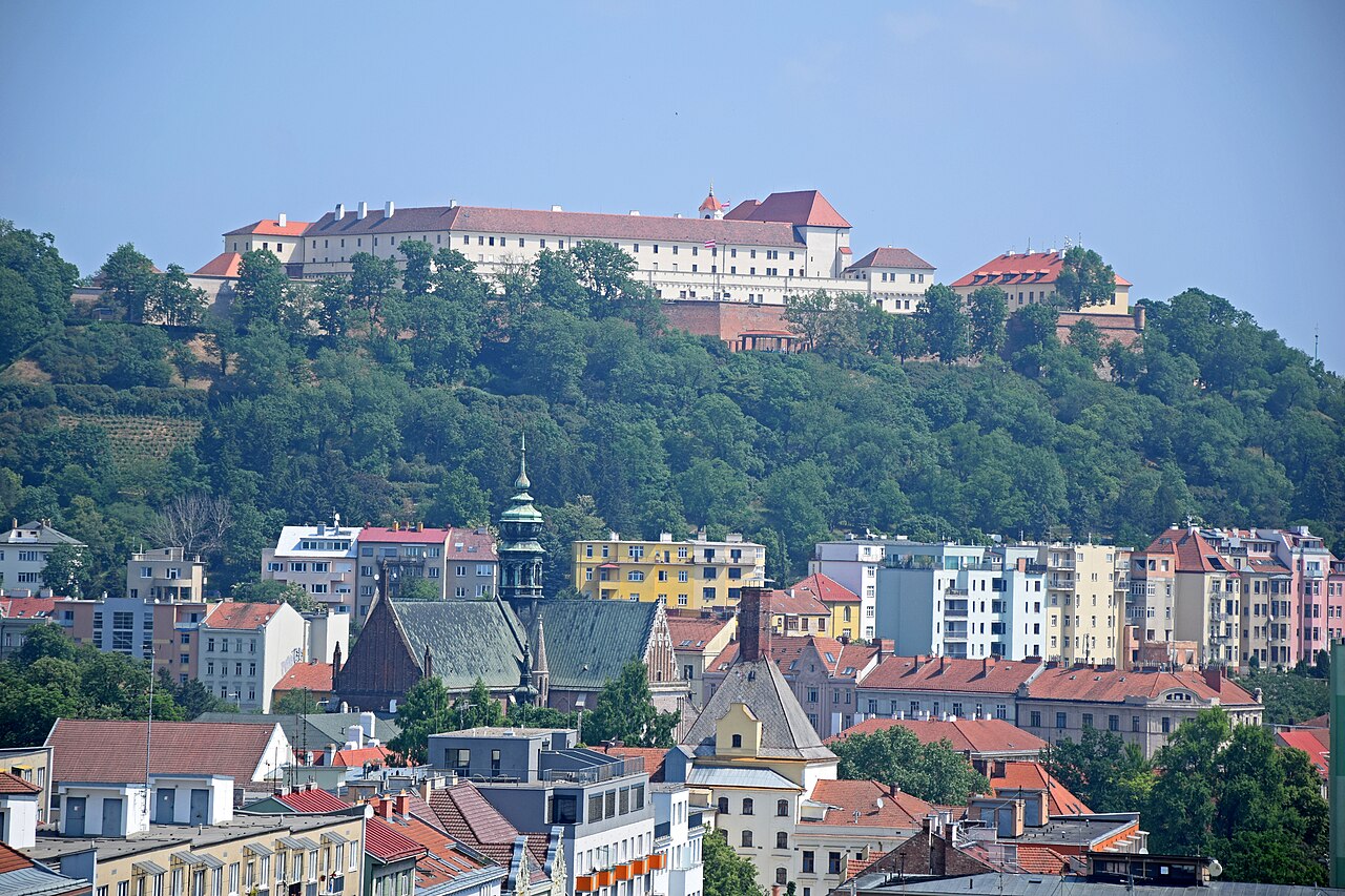 Castelo Špilberk no alto de uma colina arborizada e Basílica da Assunção de Nossa Senhora entre os edifícios urbanos abaixo.