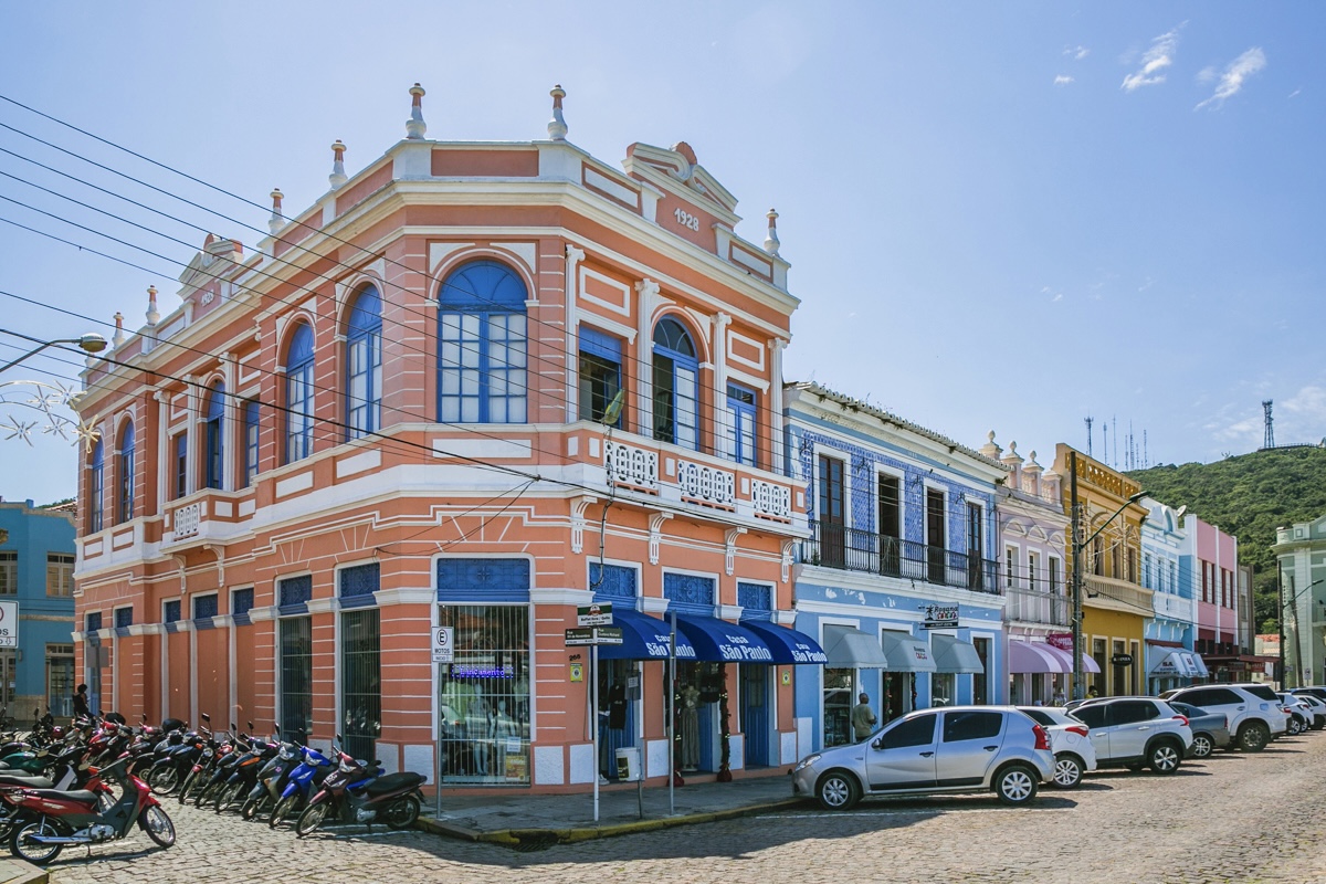 Rua de paralelepípedos com sobrados coloridos, prédio de esquina salmão com janelas azuis, veículos e morro arborizado ao fundo.