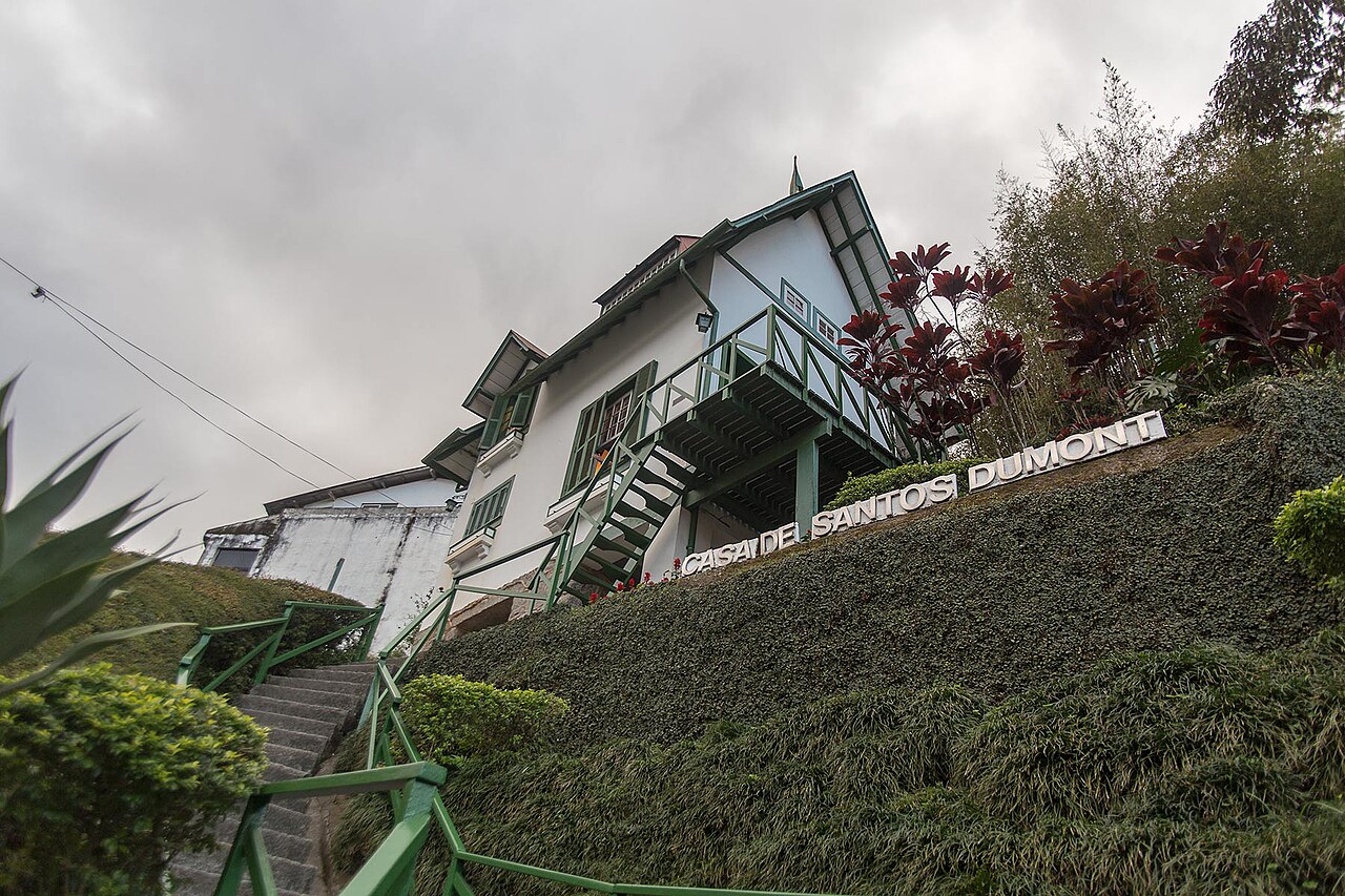 Casa de Santos Dumont branca, em terreno inclinado, com escada externa e guarda-corpos verdes, jardins e identificação diante do edifício.