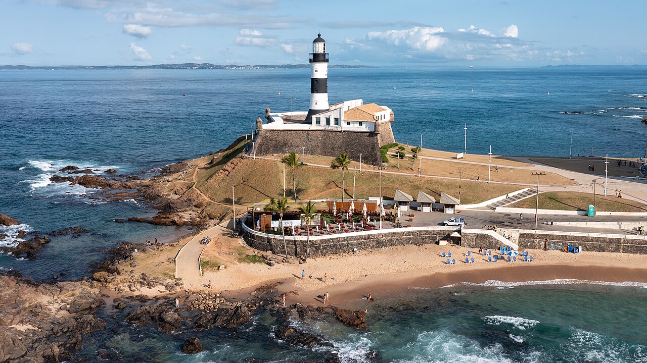 Vista elevada do Forte de Santo Antônio da Barra sobre uma ponta rochosa, com farol de faixas pretas e brancas, mar e calçadão ao redor.