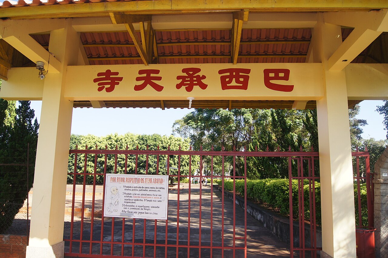 Pórtico amarelo do Templo Budista, com telhado de cerâmica, caracteres vermelhos e portão diante de um caminho arborizado.
