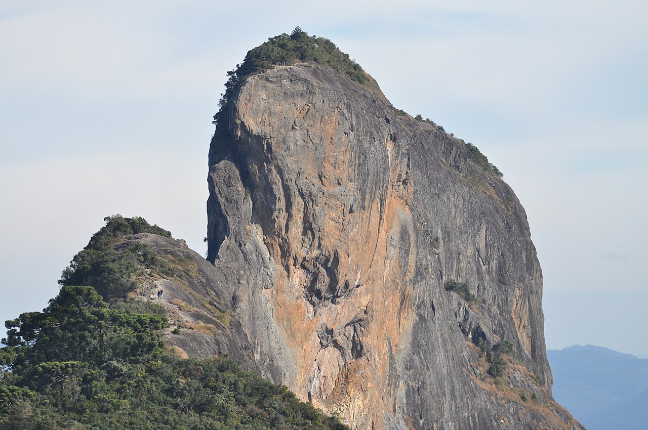 Grande paredão rochoso com marcas alaranjadas e vegetação no topo, ao lado de uma crista mais baixa com pequenas figuras humanas e montanhas ao fundo.