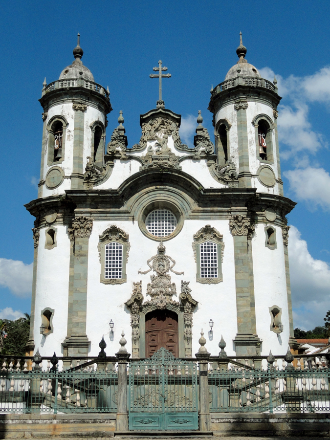 Fachada branca da Igreja de São Francisco de Assis, com ornamentação em pedra, duas torres, escadaria e portão verde.