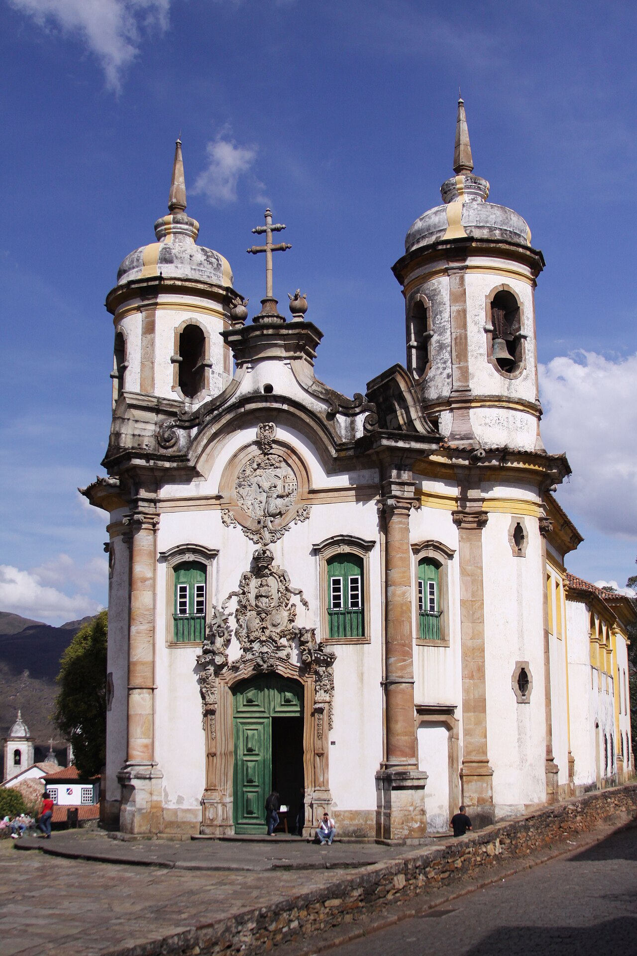 Igreja de São Francisco de Assis com fachada branca e amarela, relevos de pedra, duas torres e portas verdes, junto a calçamento em declive.