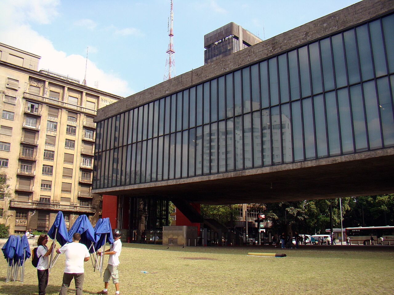 Edifício elevado de concreto e vidro do MASP, com apoios vermelhos e amplo vão livre, visto com gramado e pessoas em primeiro plano.