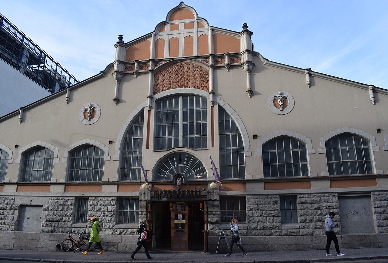 Fachada cinza e salmão do Tampere Market Hall, com janelas em arco, inscrição KAUPPAHALLI, pedestres e uma bicicleta na calçada.
