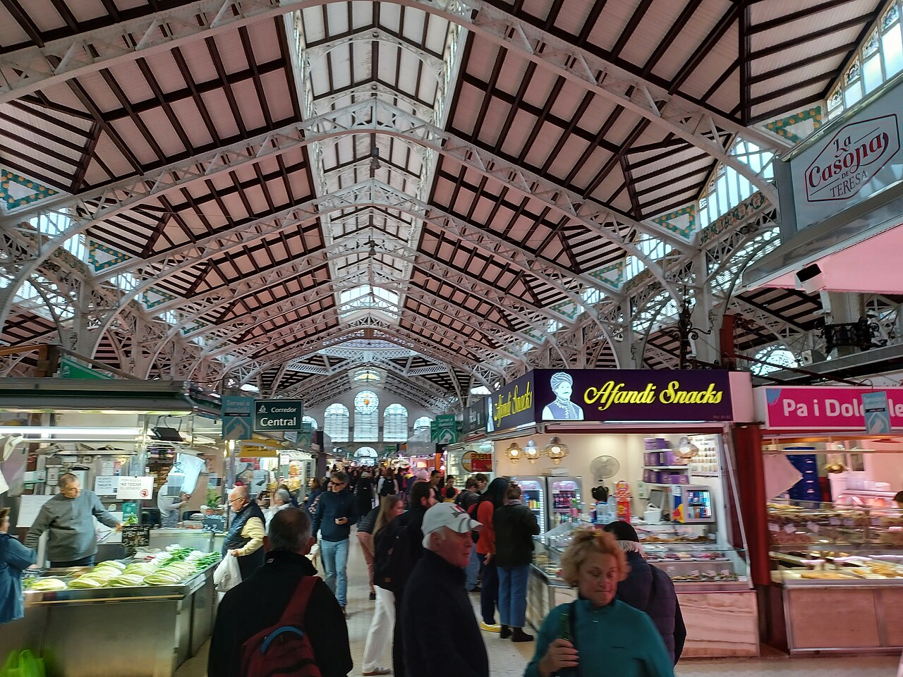 Interior do Mercado Central de Valência, com estrutura metálica clara, janelas altas e pessoas no corredor entre bancas de alimentos.