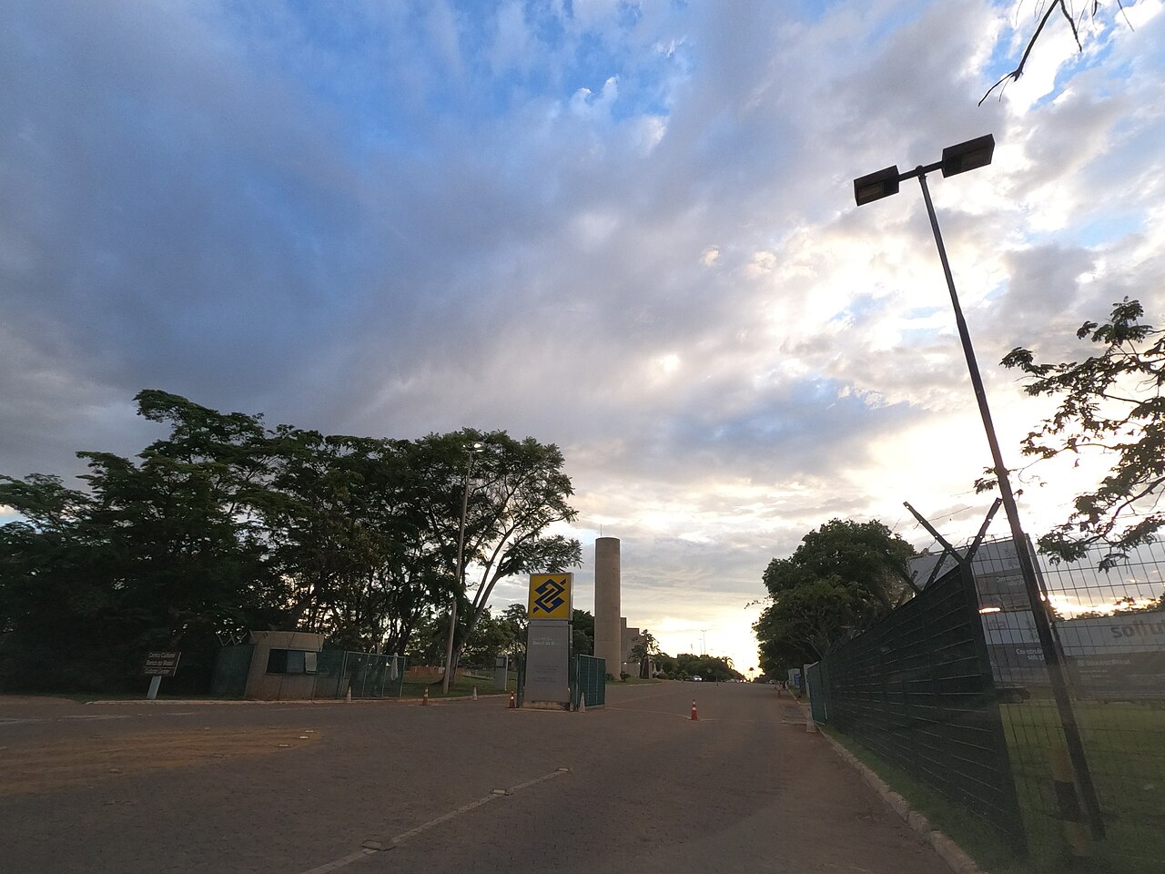 Acesso externo do CCBB com guarita, portões, grades, cones e placa do Banco do Brasil, sob céu nublado.