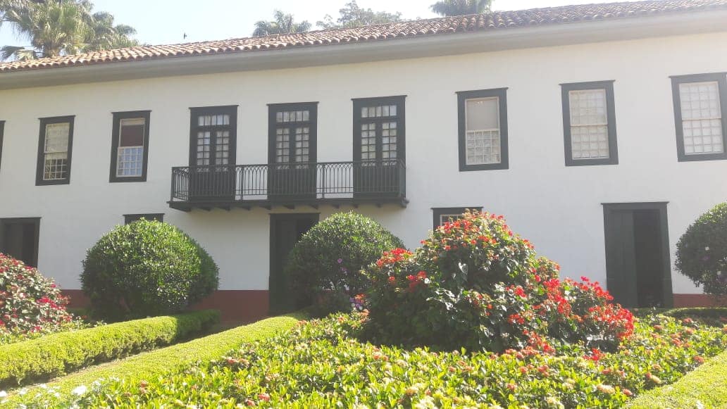 Casarão branco de dois pavimentos na Fazenda do Pinhal, com sacada central, portas e janelas escuras e jardim à frente.