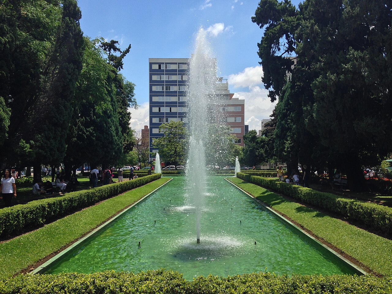 Espelho d’água com jatos de fonte na Praça da Liberdade, cercado por gramados, árvores e caminhos com pessoas.