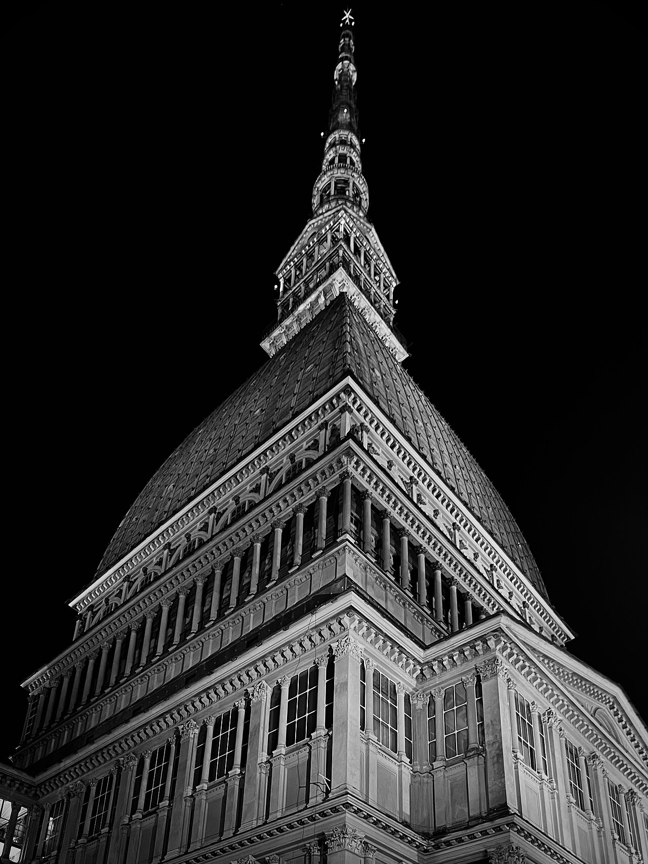 Fotografia em preto e branco da Mole Antonelliana vista de baixo, com fachada, cobertura alta e agulha iluminada contra o céu escuro.