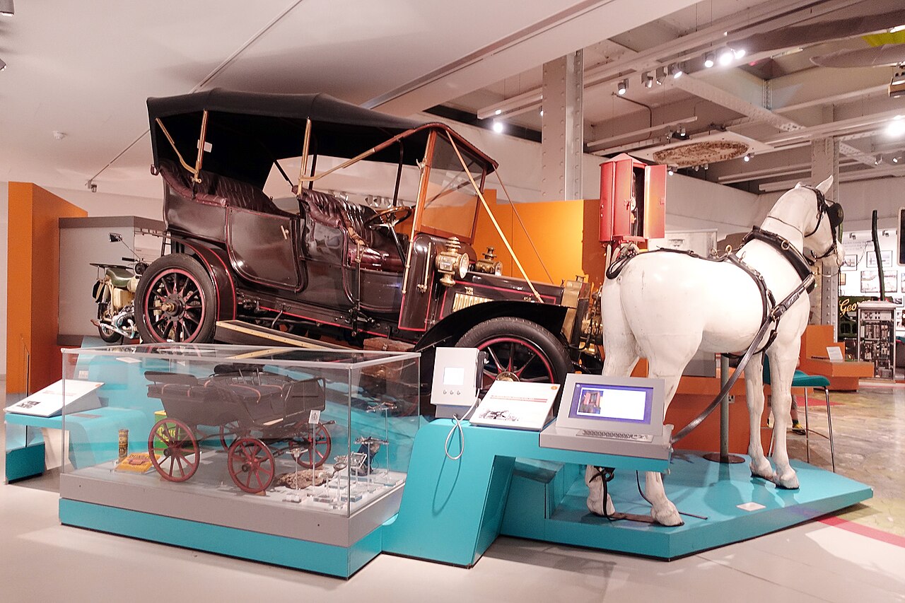 Interior do M Shed com automóvel antigo preto, réplica de cavalo branco com arreios e uma miniatura de carruagem em vitrine.