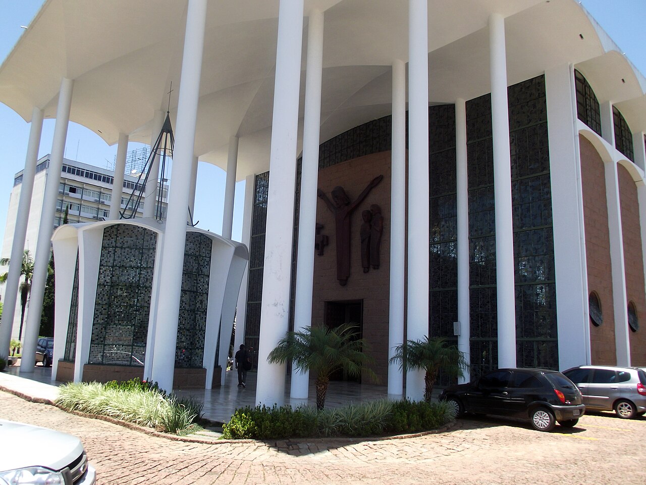 Vista externa da Catedral São Paulo Apóstolo com altos pilares brancos, cobertura ampla, painéis de vitrais escuros e relevos religiosos.