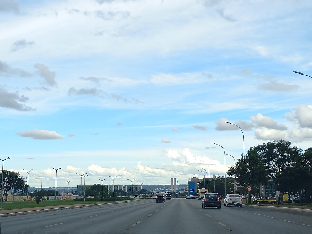Avenida larga com veículos e canteiros, sob céu com muitas nuvens, com as torres do Congresso Nacional pequenas ao fundo.