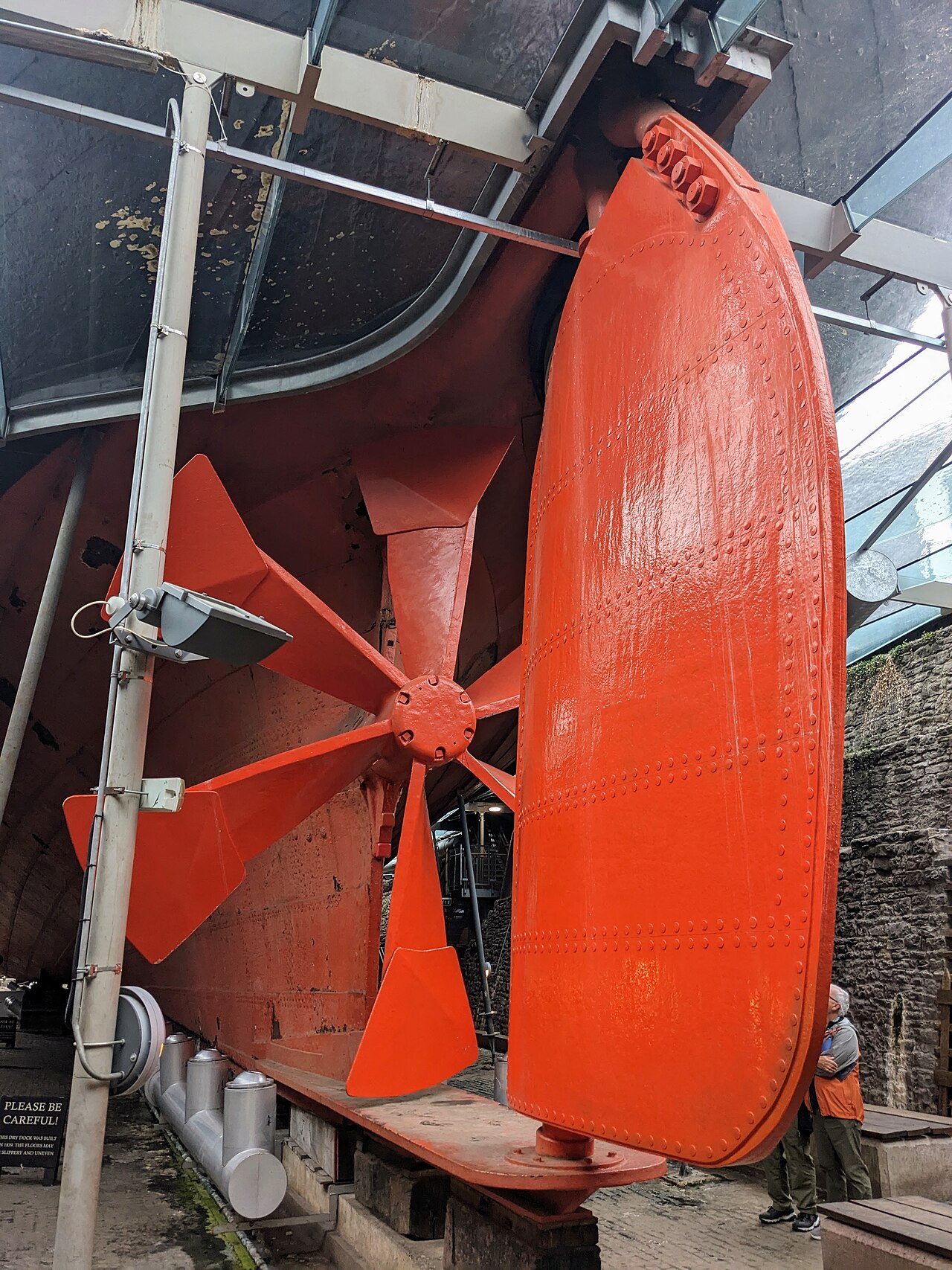 Parte inferior da popa do SS Great Britain em doca seca, com grande hélice e leme vermelho-alaranjados sob o casco.