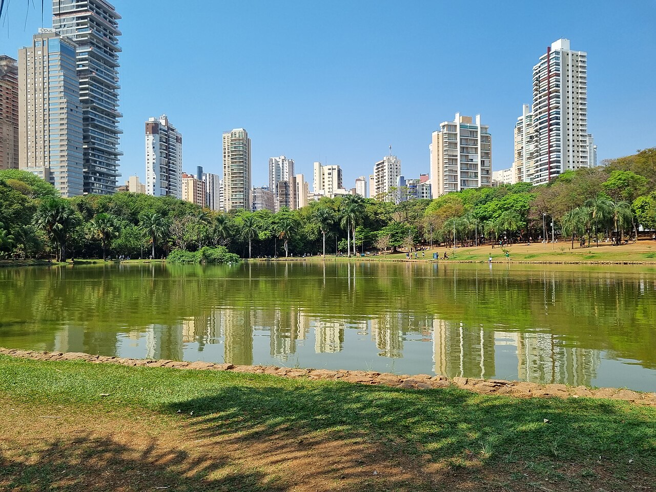 Lago do Parque Vaca Brava com reflexos de prédios altos, margens gramadas, árvores e pessoas no caminho da margem oposta.