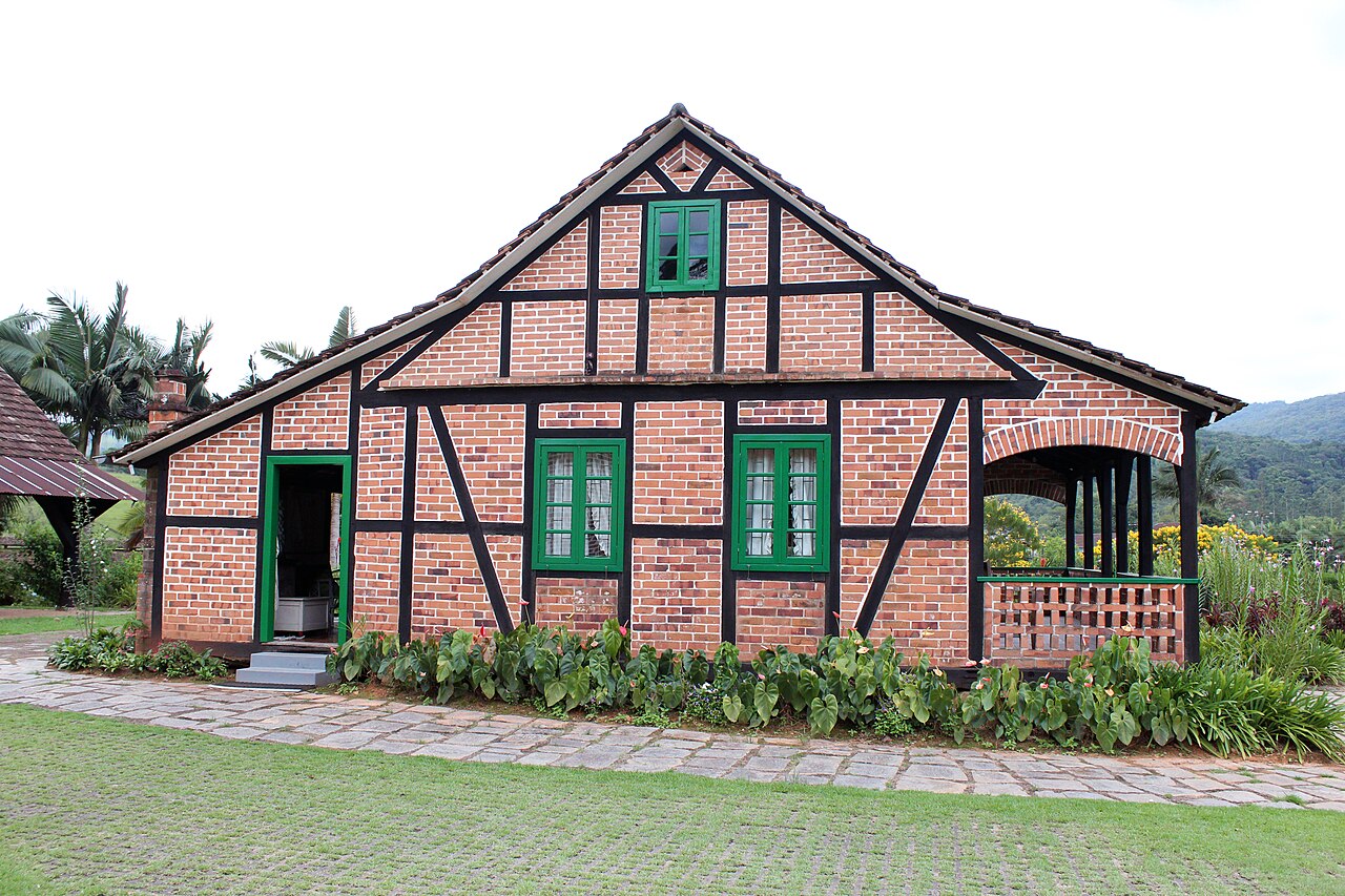Casa de tijolos e madeira escura em enxaimel, com portas e janelas verdes, varanda lateral e jardim.