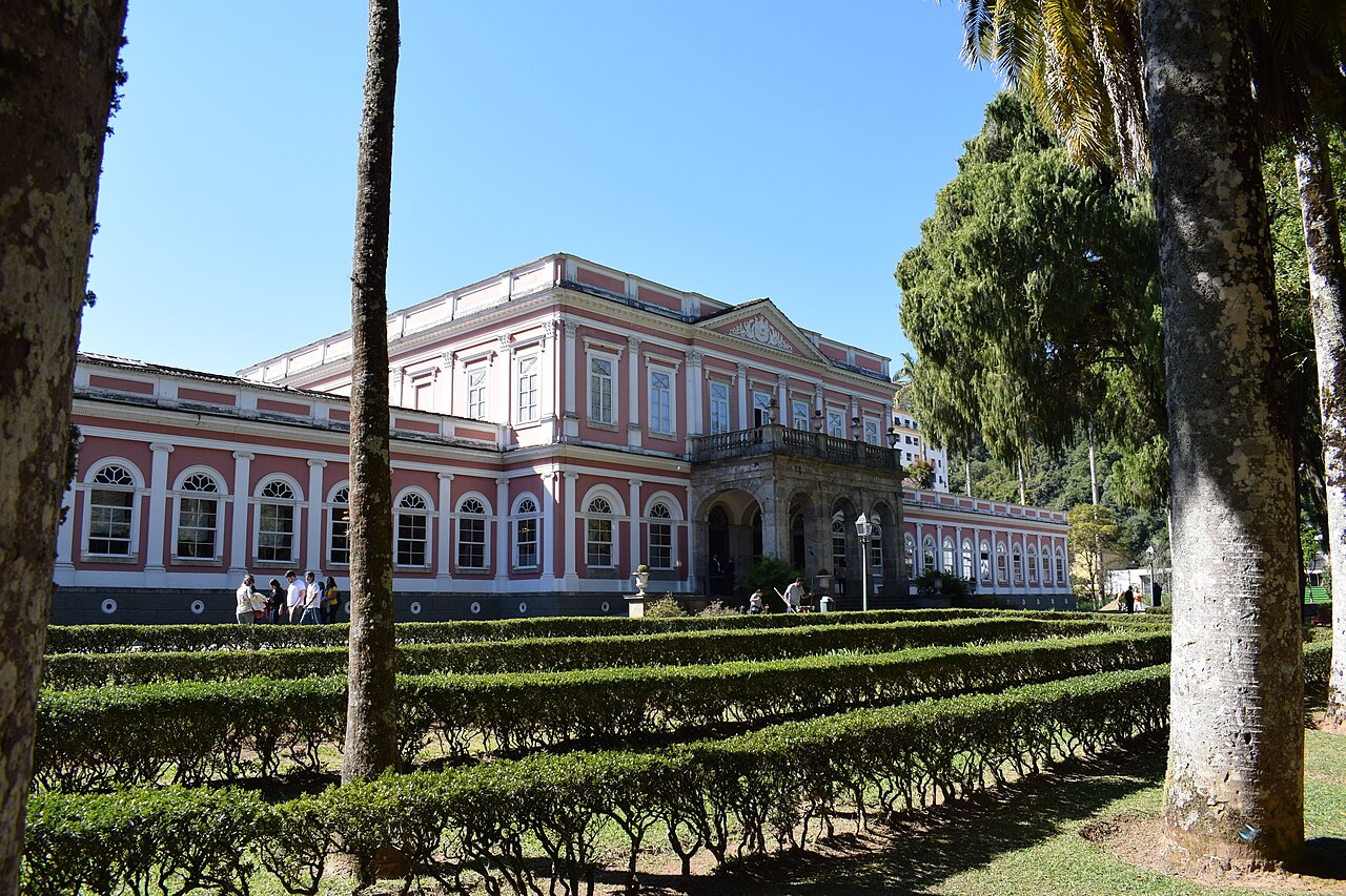 Museu Imperial visto em ângulo, com fachada rosa e branca, pórtico de pedra, sebes geométricas, palmeiras e visitantes nos jardins.