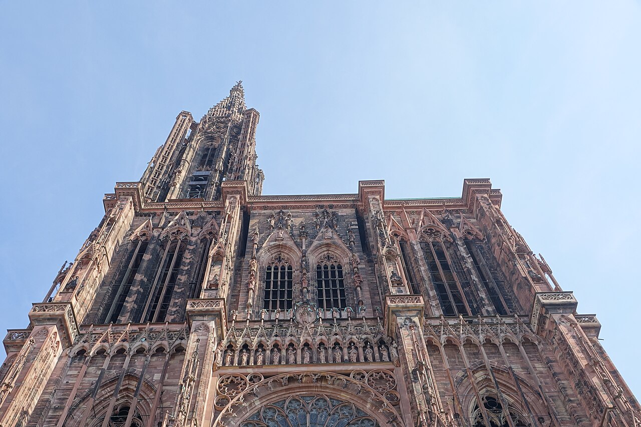 Fachada gótica de pedra rosada da Catedral Notre-Dame de Estrasburgo vista de baixo, com esculturas, janelas altas e torre sob céu azul.