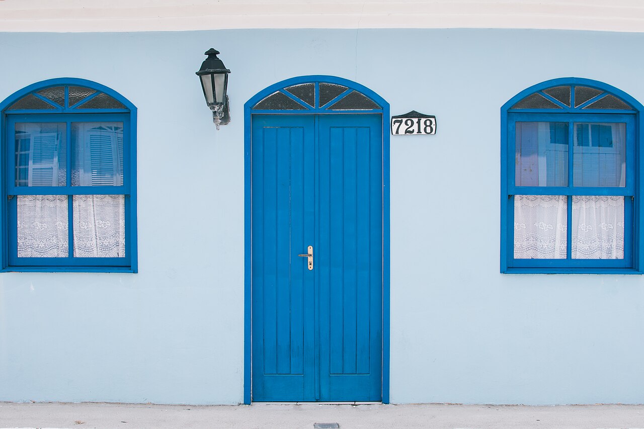Fachada azul-clara no Ribeirão da Ilha, com porta e duas janelas azuis, cortinas rendadas e luminária preta.