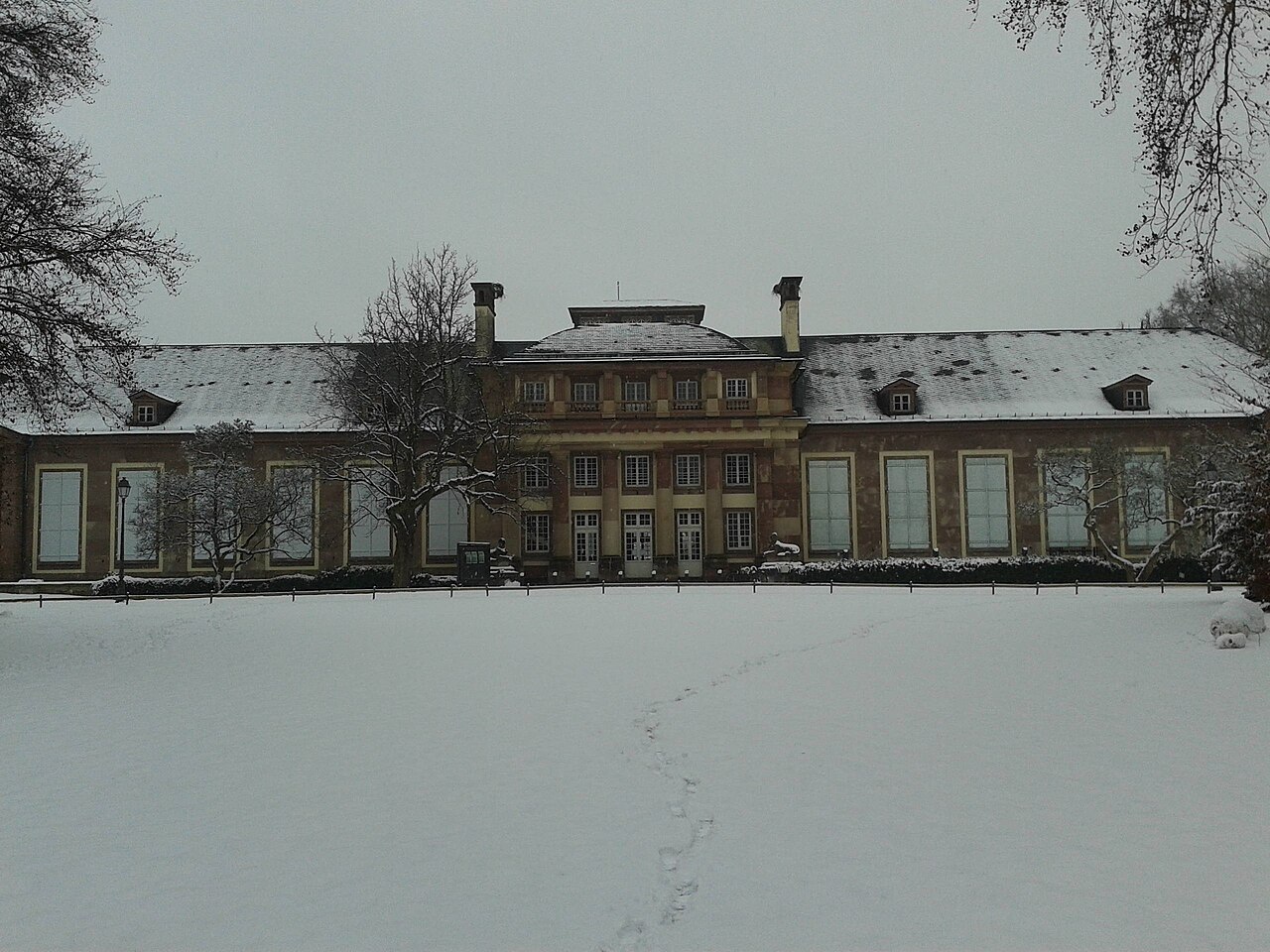 Pavillon Joséphine no Parc de l’Orangerie, com telhado e gramado cobertos de neve, pegadas e árvores sem folhas.
