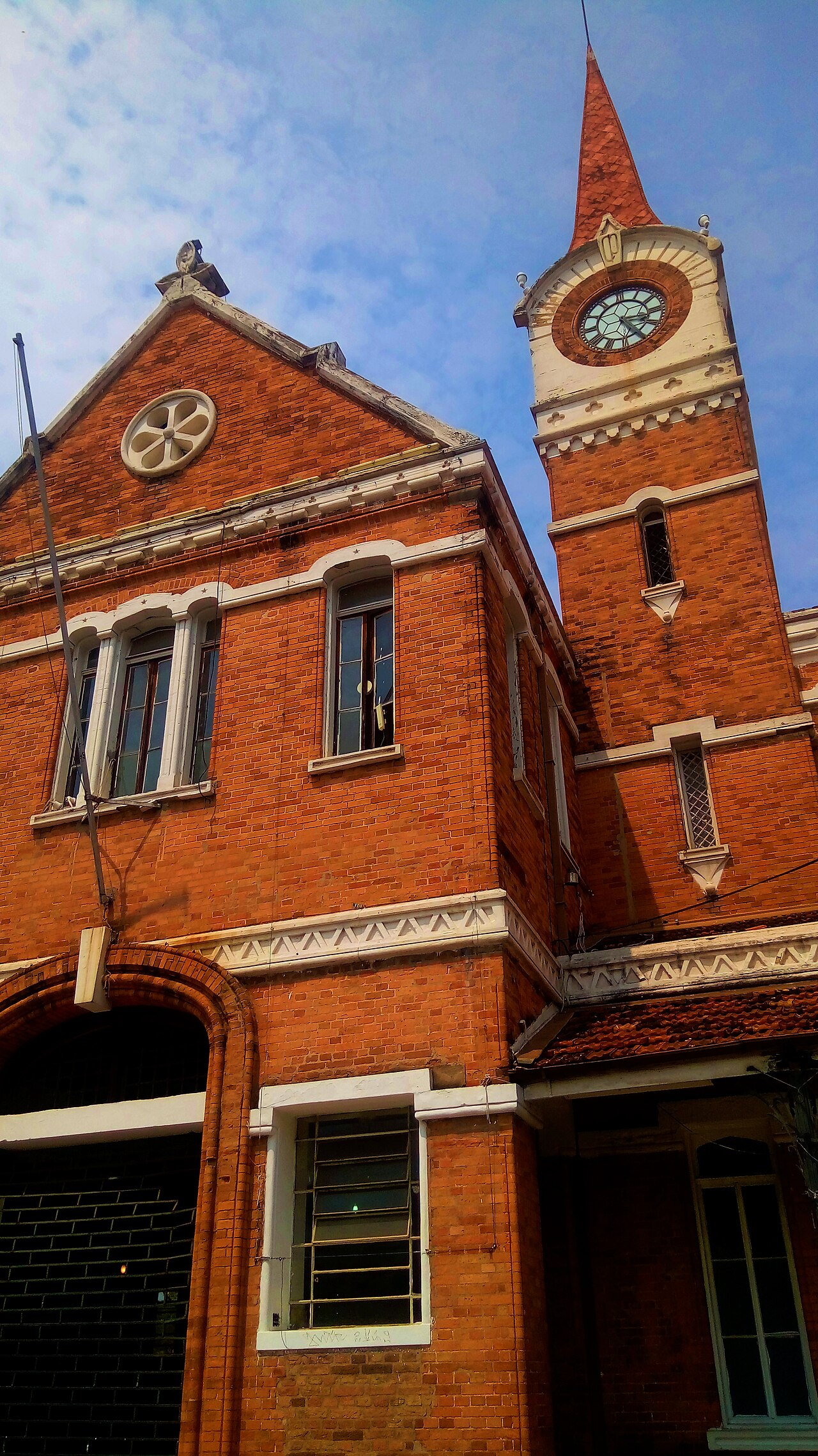 Enquadramento parcial de baixo para cima da Estação Cultura, com fachada de tijolos vermelhos, detalhes claros e torre com relógio sob céu azul.