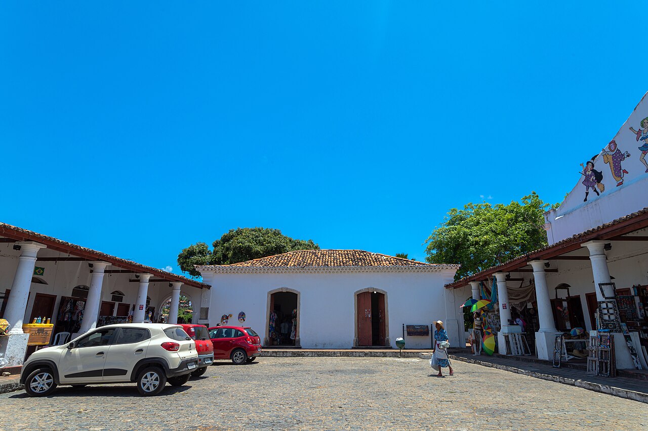 Pátio de pedras do Mercado da Ribeira cercado por construções brancas com colunas e telhados cerâmicos, com carros à esquerda, uma pessoa à direita e objetos coloridos nas laterais.