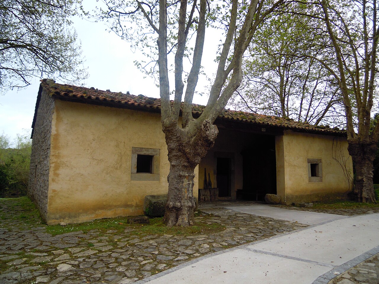 Casa térrea amarelada com lateral de pedra, telhado cerâmico, duas pequenas janelas e entrada central recuada, com árvores diante da fachada.