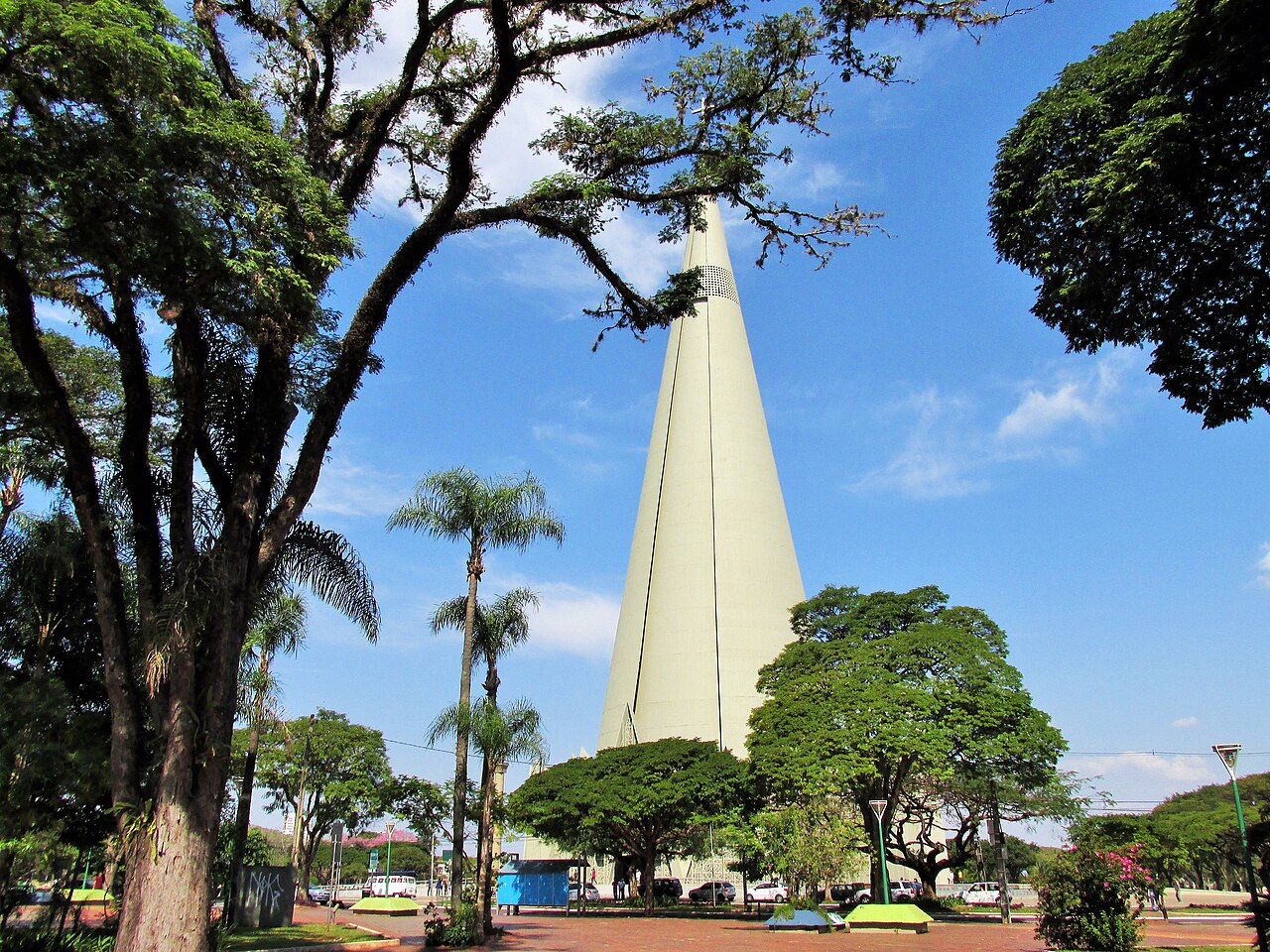 Catedral de Maringá com construção alta e cônica parcialmente encoberta por árvores, palmeiras e praça pavimentada sob céu azul.