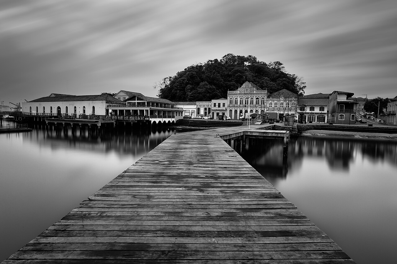 Fotografia em preto e branco com píer de madeira, água com reflexos, casario à beira d’água e morro arborizado ao fundo.