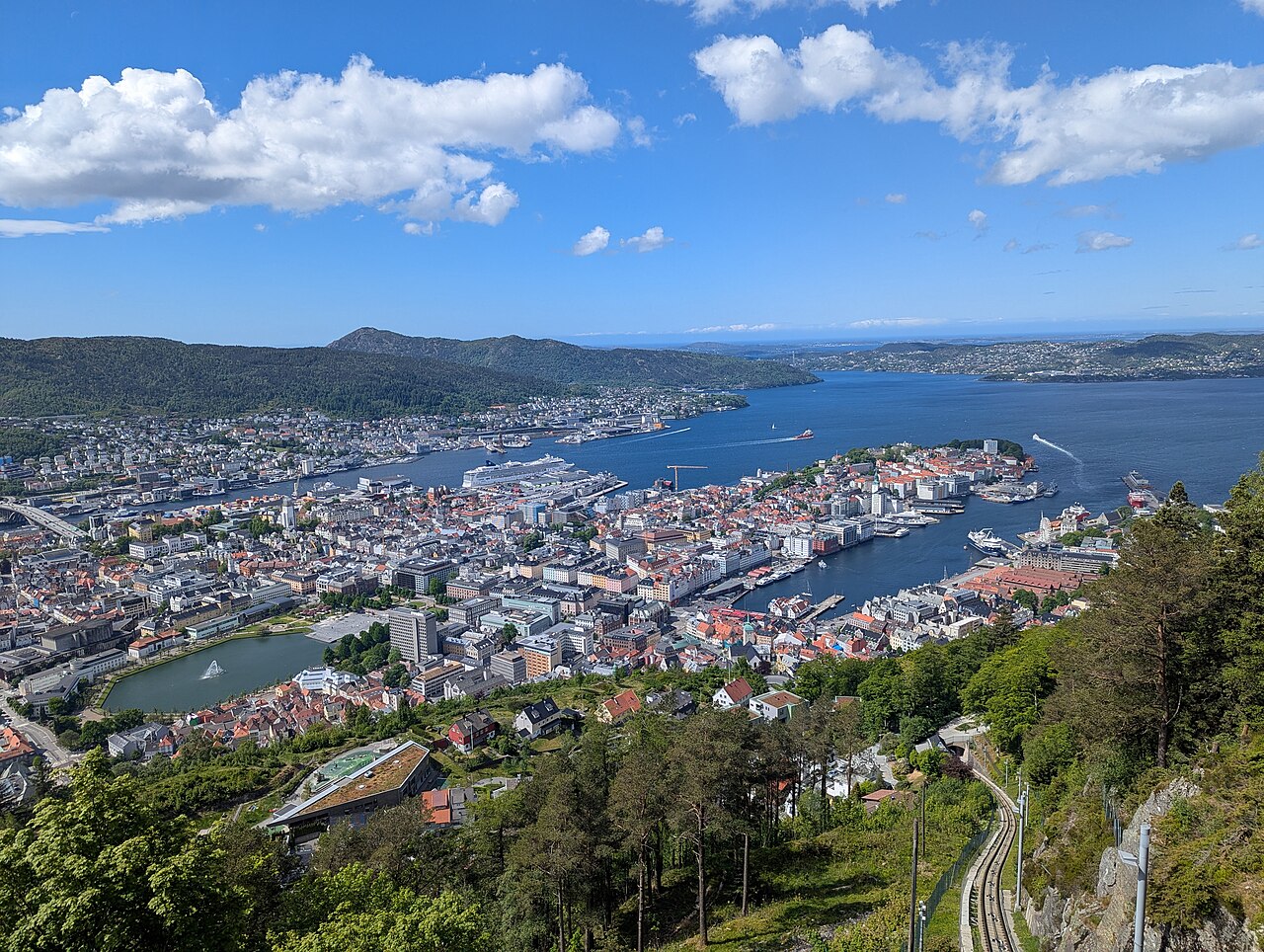 Vista elevada de Bergen com porto, casario, lago e morros arborizados, com trilhos de funicular à direita.