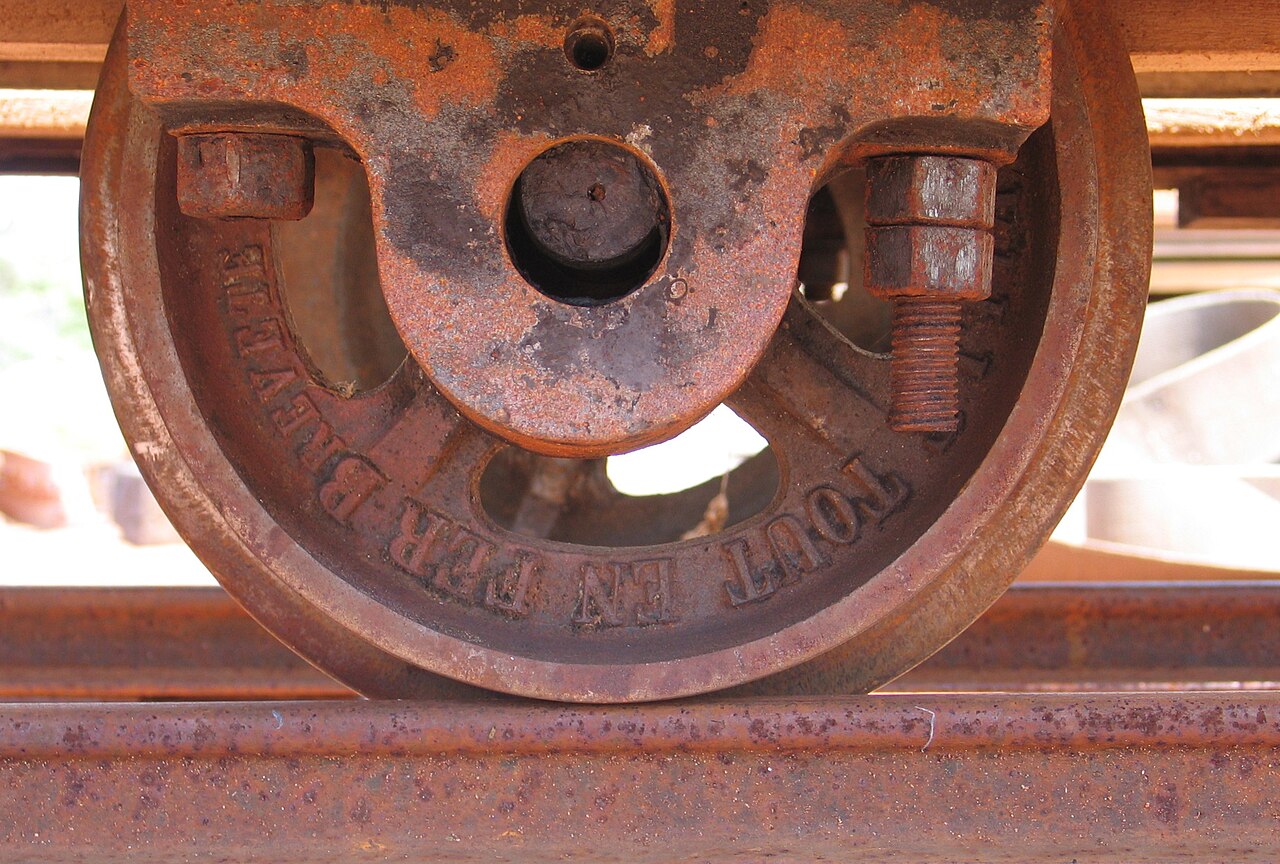 Detalhe de roda metálica enferrujada sobre um trilho, com eixo, suporte, porcas e parafuso aparente.