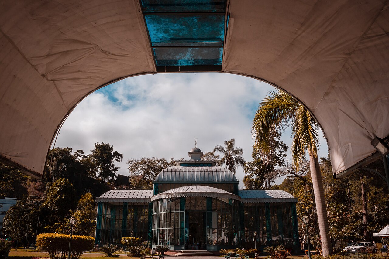 Palácio de Cristal visto de frente, com estrutura verde e vidro, cercado por jardins e enquadrado por uma grande cobertura clara em arco.