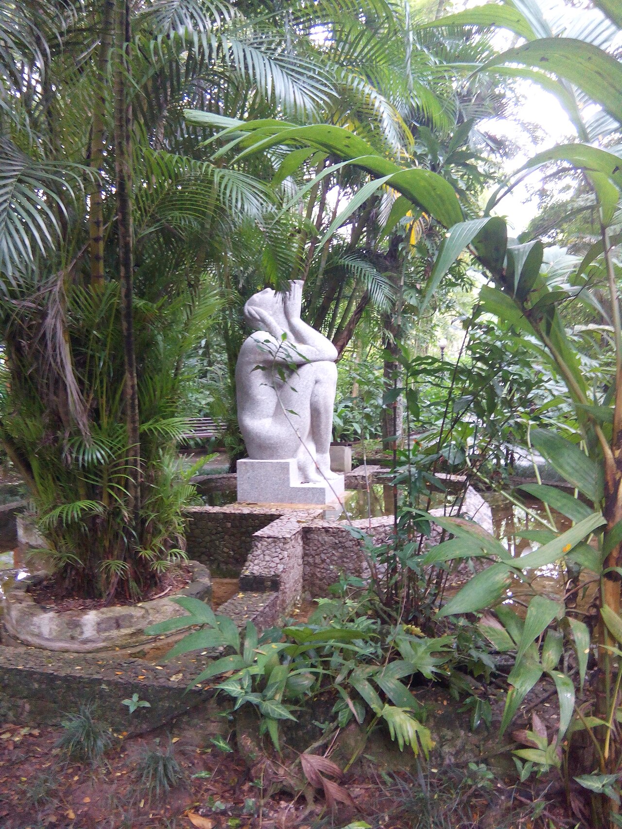 Escultura clara de figura humana sentada sobre pedestal, cercada por palmeiras, vegetação, pequenos espelhos d’água e um banco ao fundo.