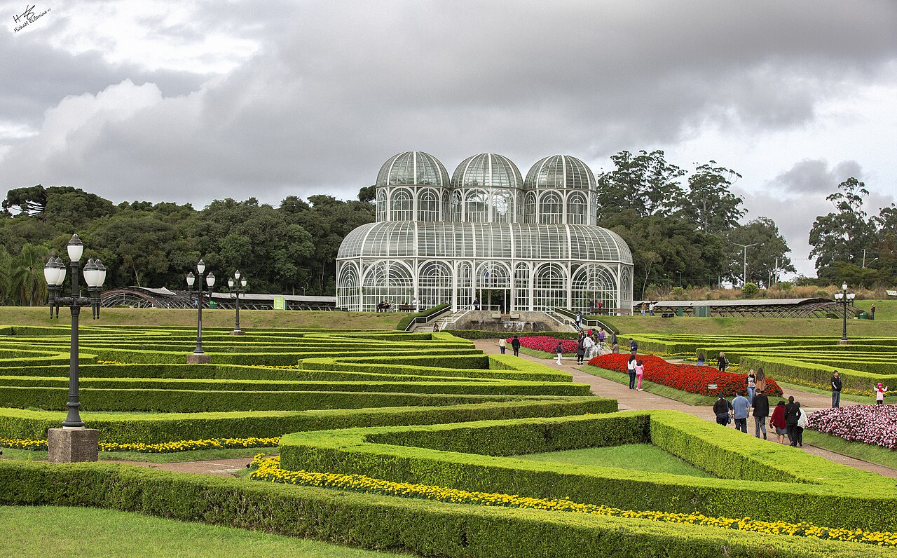 Estufa de vidro com estrutura branca e três cúpulas, diante de jardins geométricos, canteiros floridos e caminhos com visitantes.