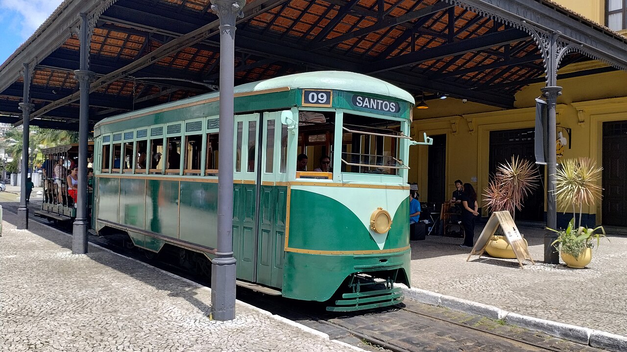 Bonde verde e creme com número 09 e letreiro Santos sobre trilhos sob cobertura metálica, com pessoas e edifício amarelo ao fundo.