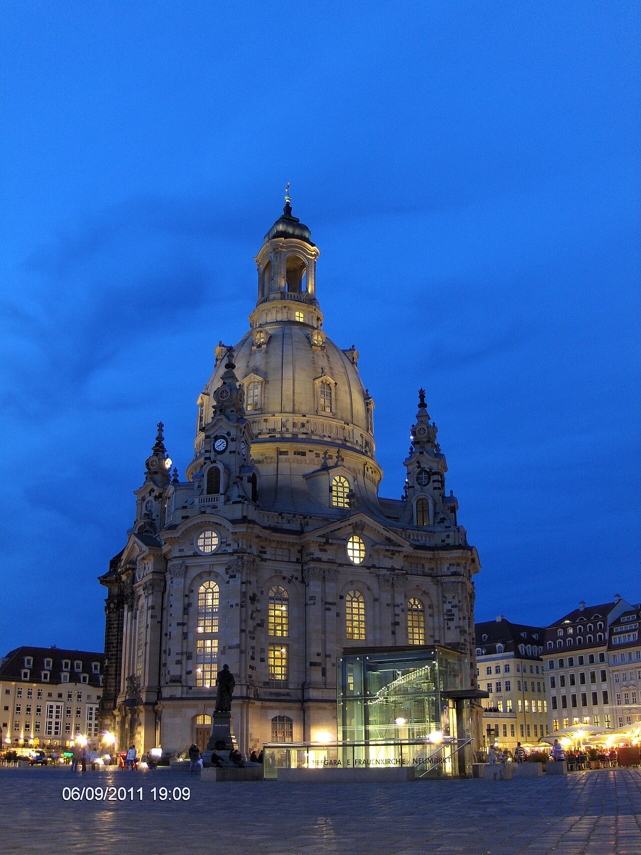 Fachada de pedra clara e grande cúpula da Frauenkirche, com pedestres na praça ao entardecer.