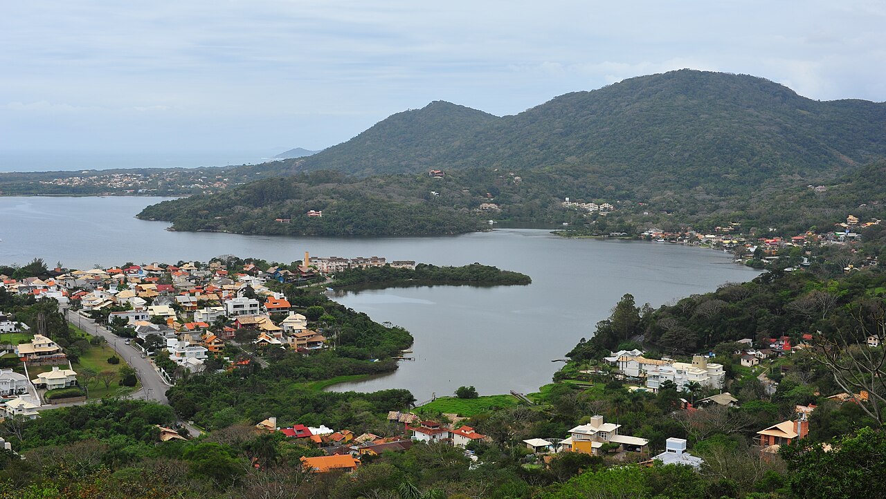 Vista elevada da Lagoa da Conceição, com casas e vegetação em primeiro plano e morros ao fundo sob céu nublado.