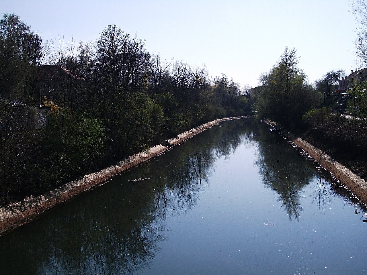 Águas do Karl-Heine-Kanal refletindo árvores, com margens revestidas, vegetação e um caminho com escadas à direita.