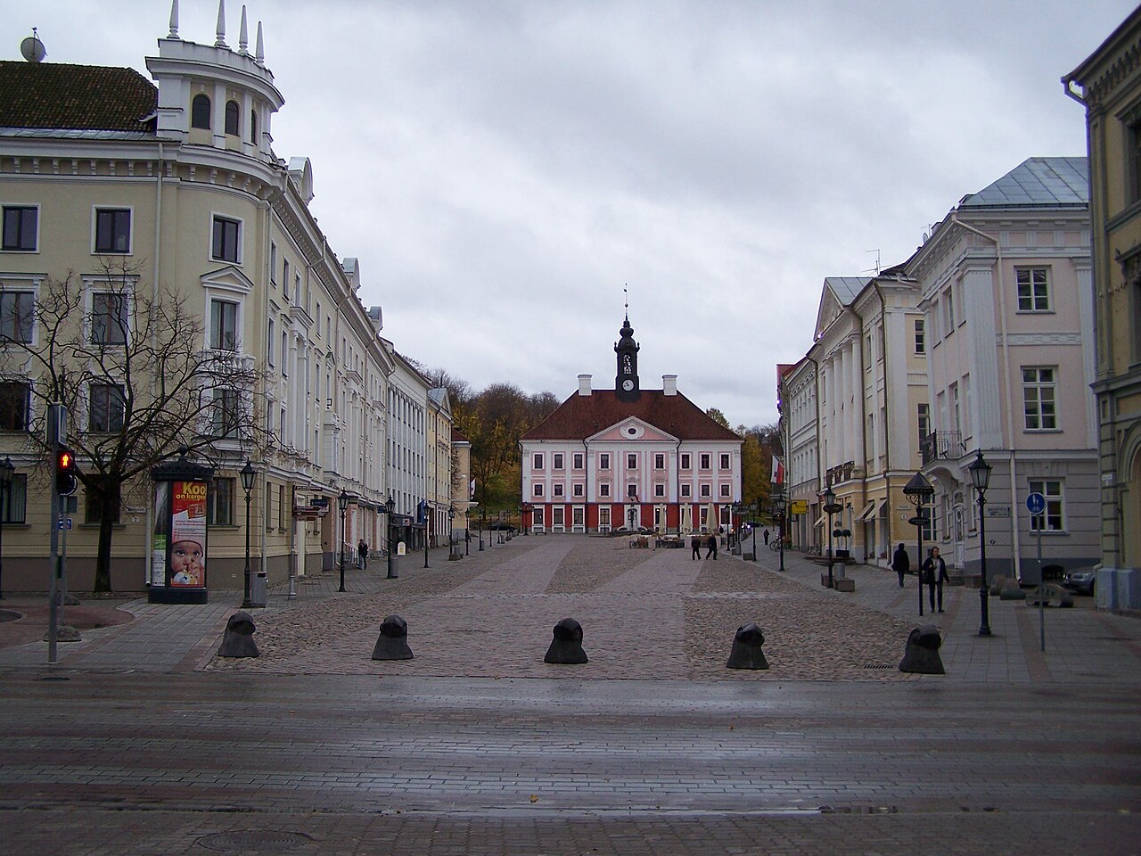 Praça de pedras ladeada por edifícios claros, com a prefeitura rosada de torre com relógio ao fundo, pedestres e céu encoberto.