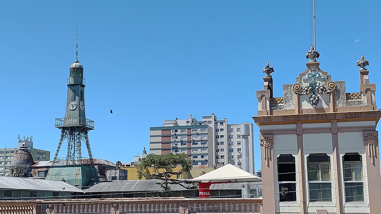 Telhados do Mercado Público de Pelotas, com torre metálica de relógio à esquerda, fachada clara ornamentada à direita e prédios ao fundo.