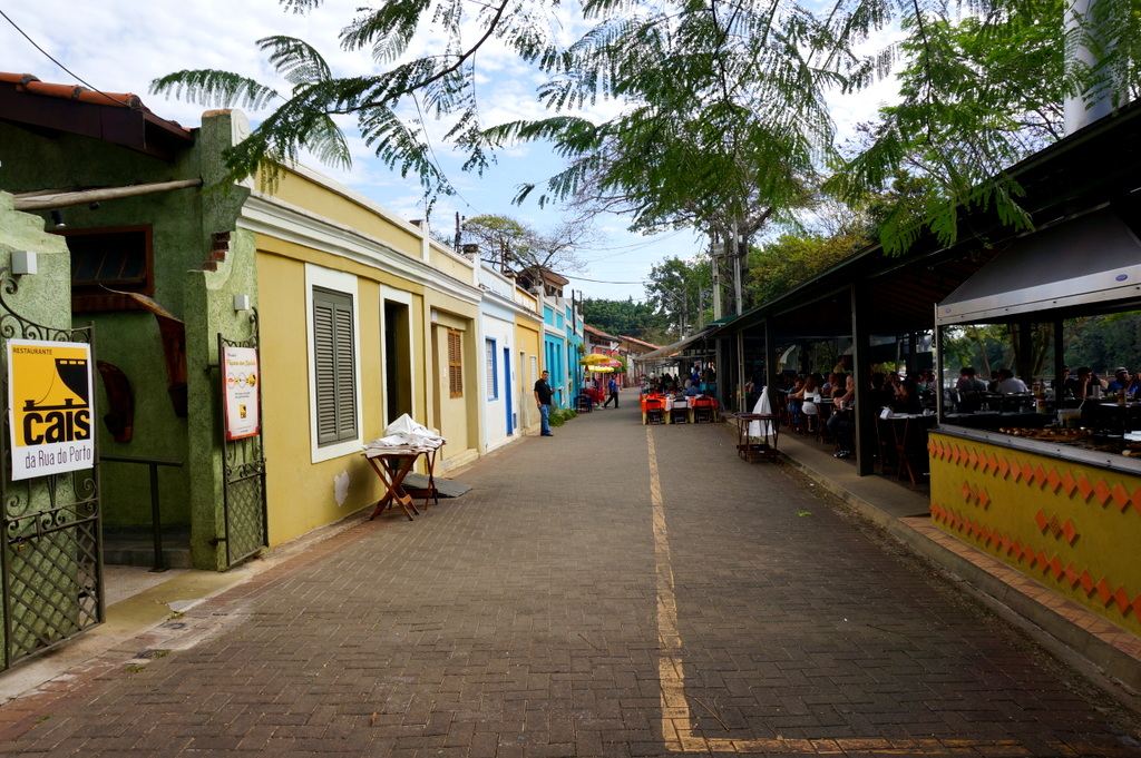 Rua pavimentada com tijolos, casas térreas coloridas à esquerda e mesas e cadeiras em áreas cobertas à direita, sob árvores.