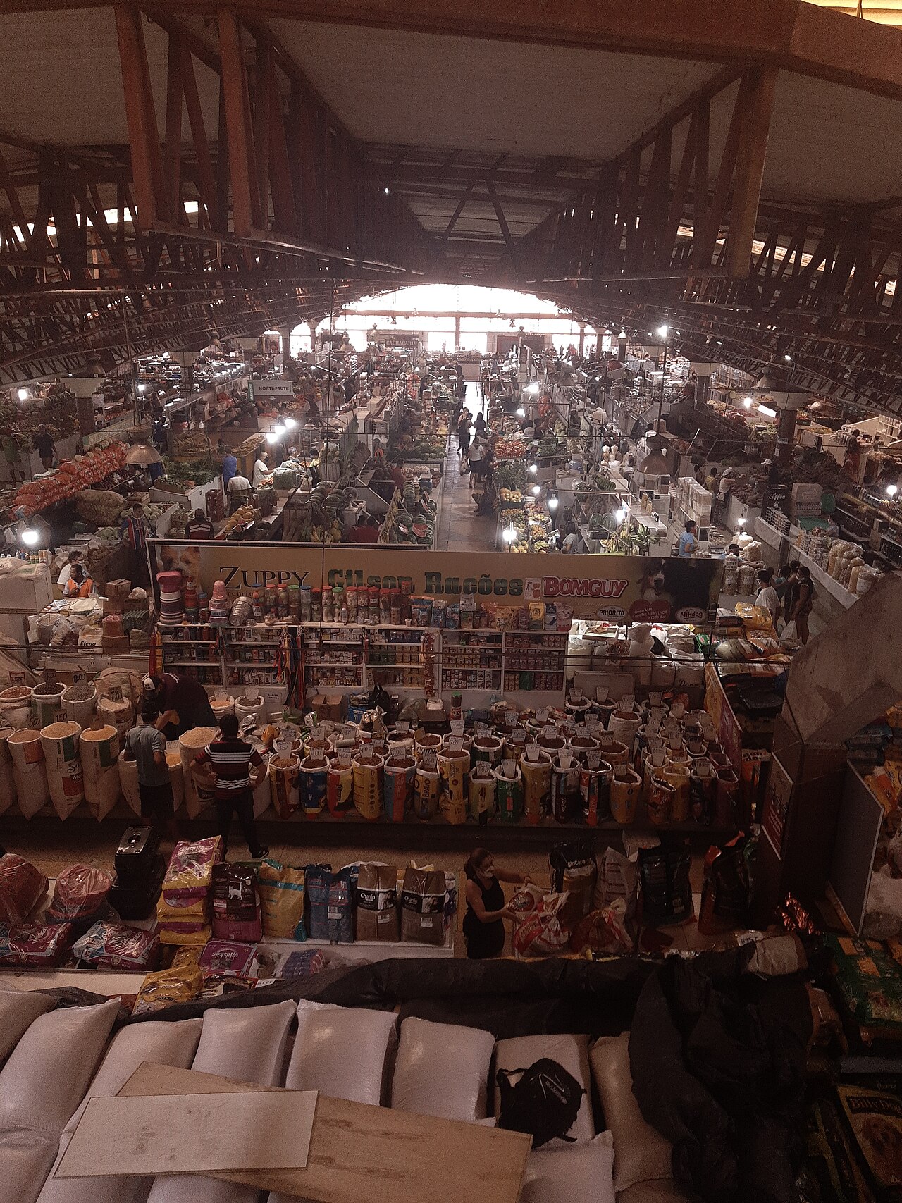 Vista elevada de mercado coberto com estrutura metálica, corredores entre bancas, pessoas, sacos de mercadorias a granel e produtos embalados.