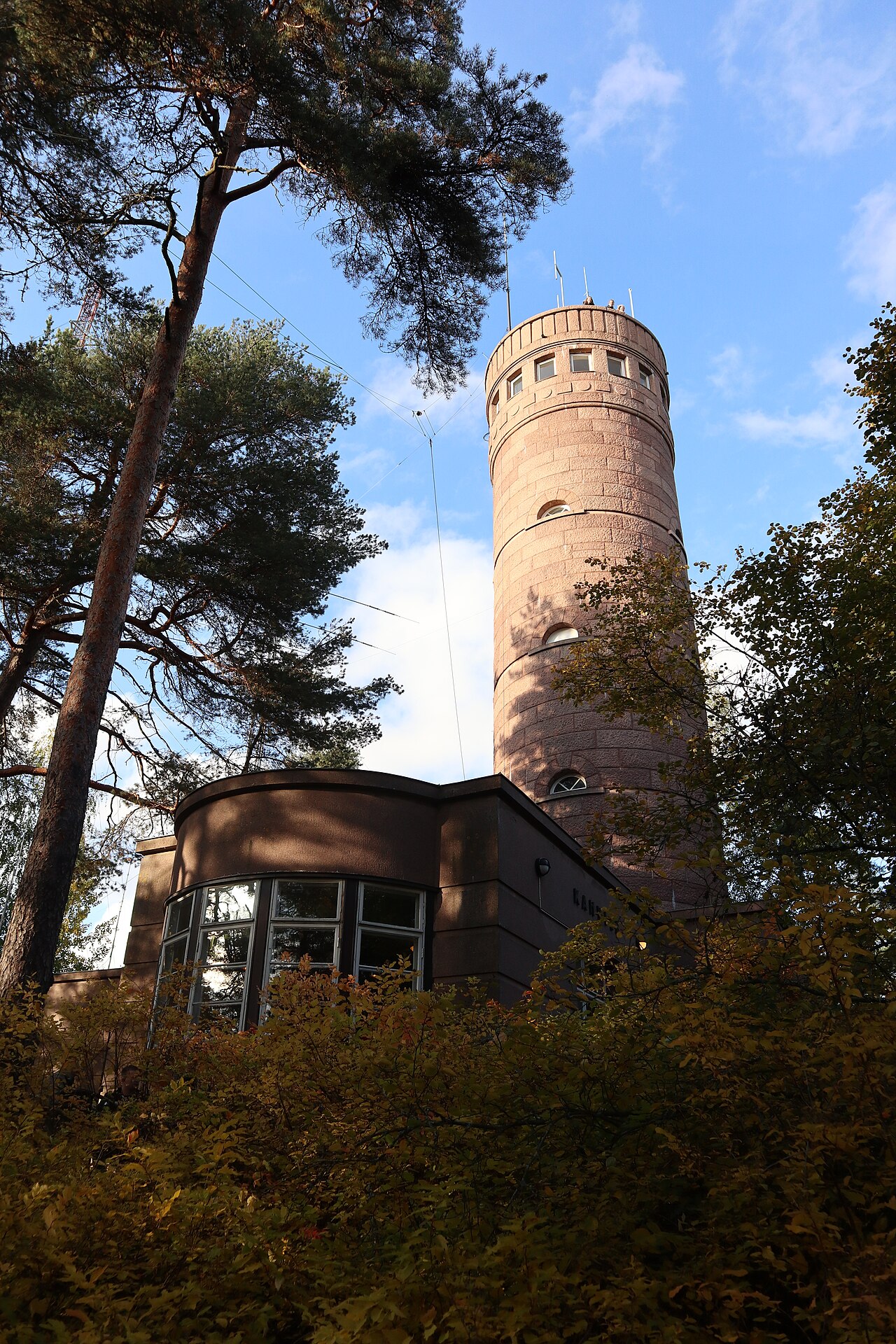 Torre cilíndrica de pedra rosada de Pyynikki vista de baixo, com árvores de folhagem amarelada e céu azul.