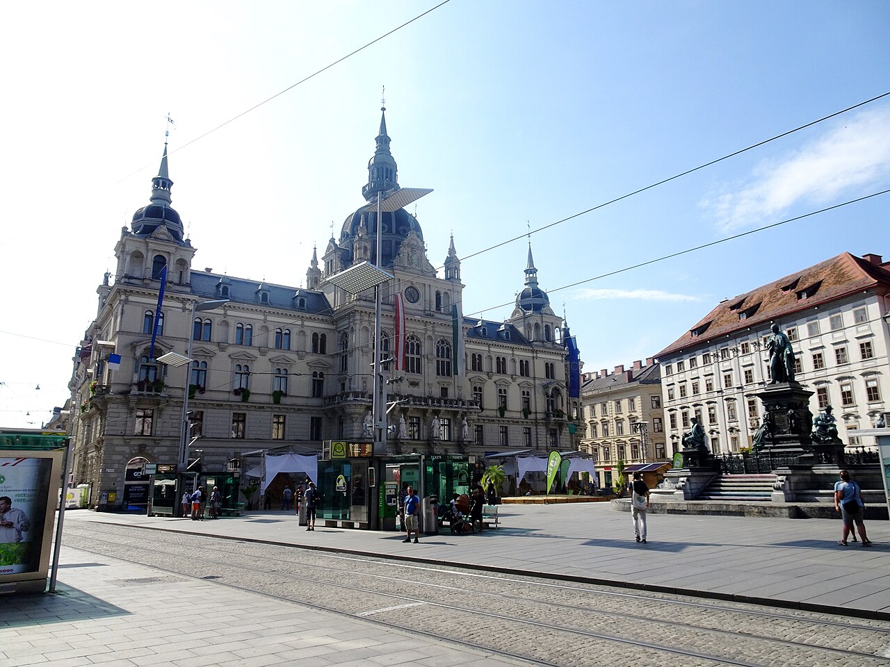 Praça pavimentada com trilhos de bonde, pedestres e quiosques diante da prefeitura de Graz, de fachada ornamentada e grande cúpula.
