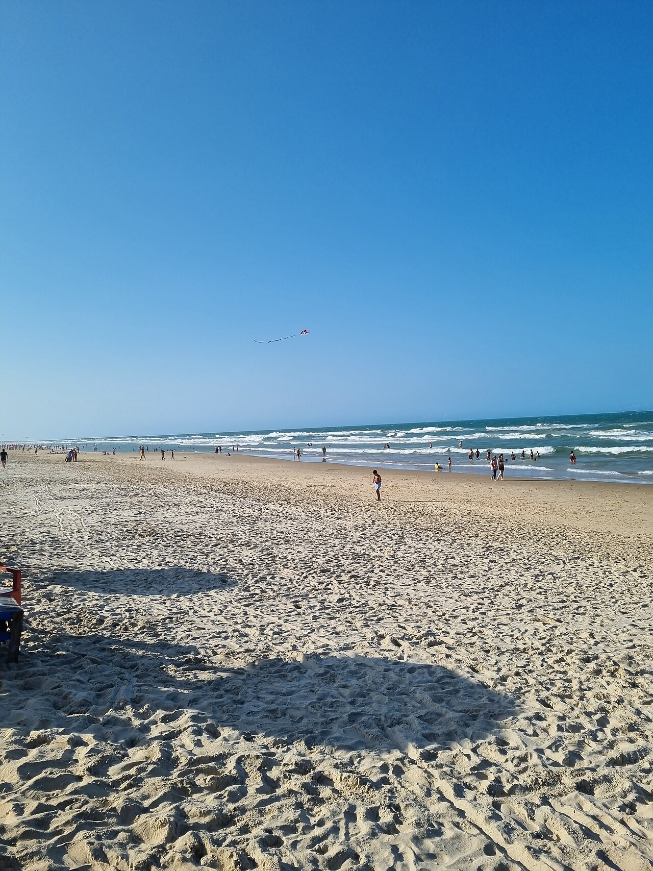 Praia do Futuro com faixa ampla de areia, banhistas, ondas com espuma e uma pipa no céu azul.