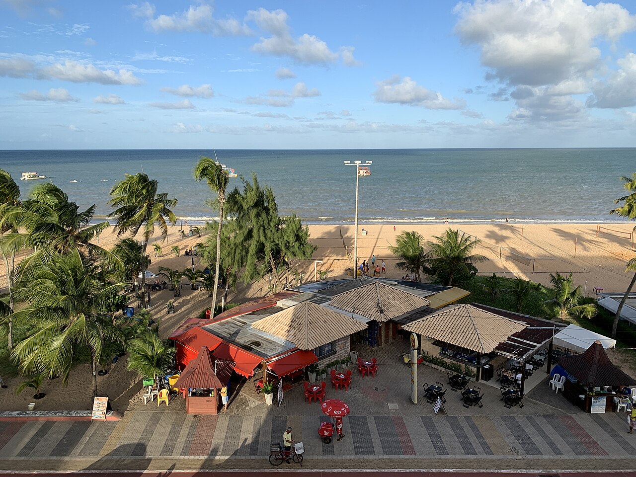 Vista elevada da Praia de Tambaú com areia, coqueiros, redes esportivas, pequenas embarcações, quiosques e ciclovia.