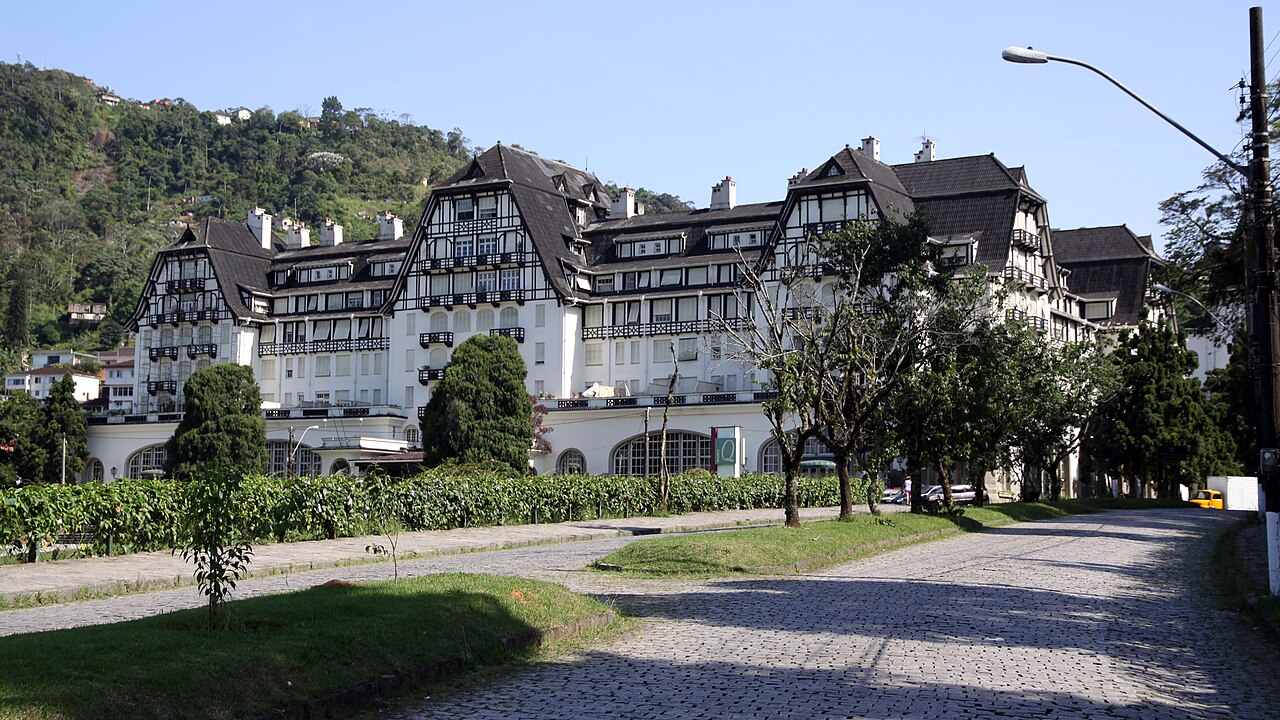Edifício do Quitandinha com fachadas brancas e elementos escuros, telhados altos, jardins, via de paralelepípedos e morro arborizado ao fundo.