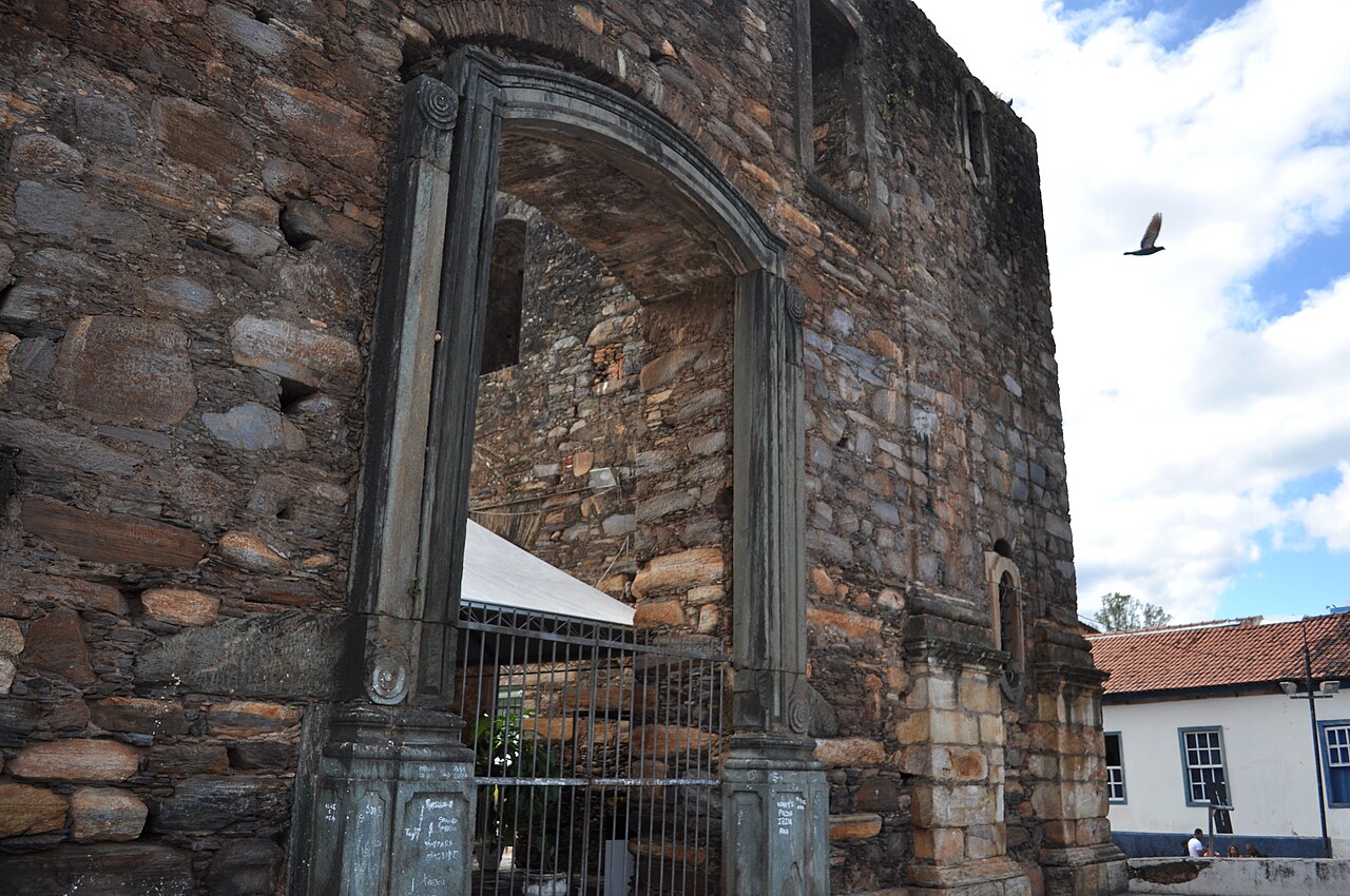 Paredes altas de pedra aparente e portal em arco com grades na construção inacabada da Igreja do Rosário.