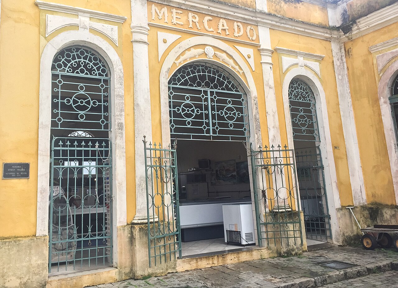 Fachada amarela do Mercado com ornamentos brancos, três vãos em arco, grades verdes e calçamento de pedra.